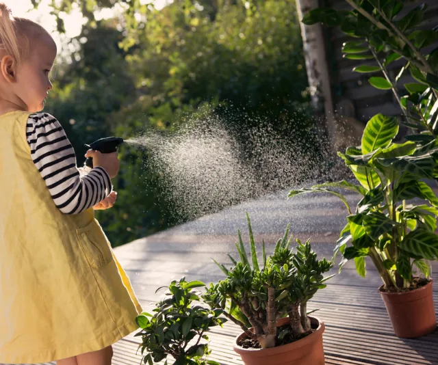 Child in a yellow dress waters potted plants with a spray bottle on a sunny deck.