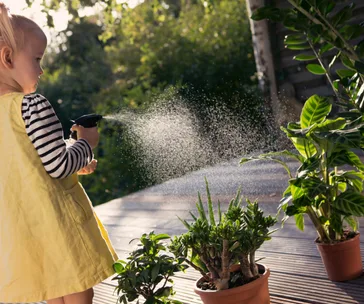 Child in a yellow dress waters potted plants with a spray bottle on a sunny deck.