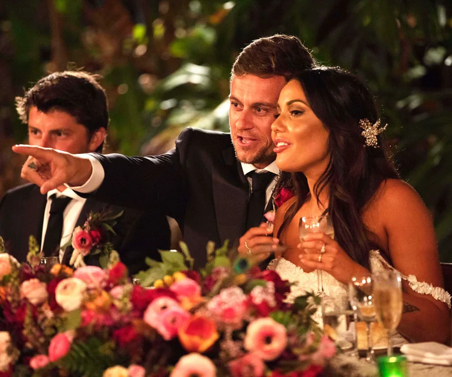 A couple in formal attire sitting at a flower-adorned table, the man points while both look at something off-camera.