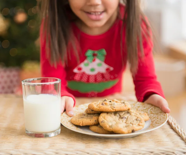 Girl holding plate of cookies with glass of milk, wearing red festive shirt.