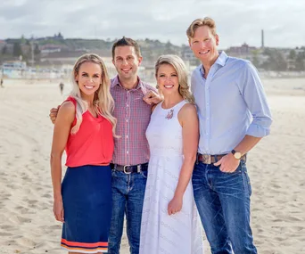 Four people stand smiling on a sunny beach, dressed casually.