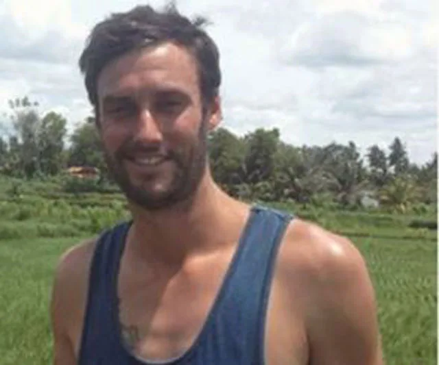 A person in a sleeveless top stands outdoors with lush greenery and a cloudy sky in the background.