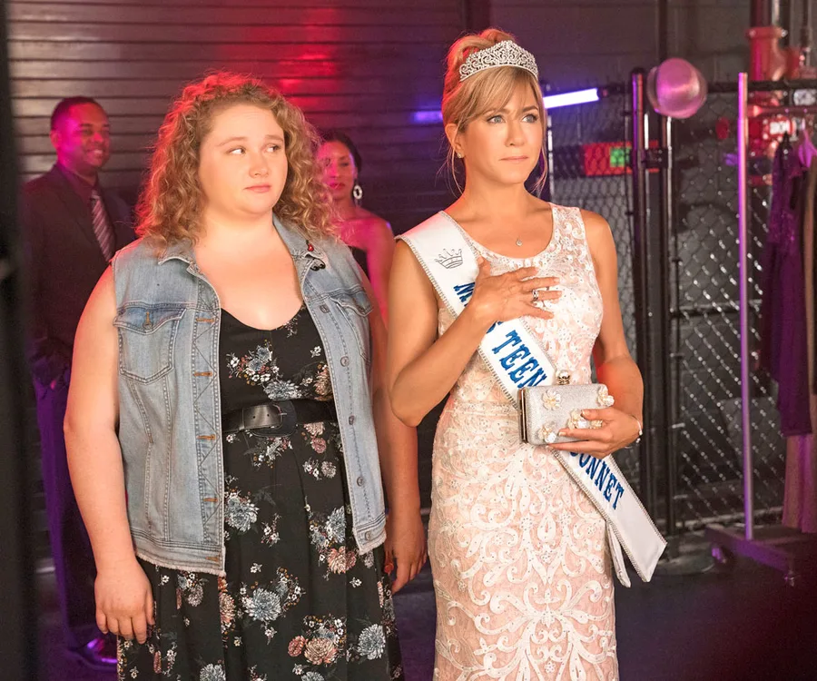 Mother and daughter stand together; daughter in casual attire, mother in pageant dress and sash, touching her chest.