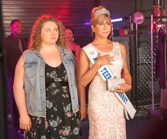 Mother and daughter stand together; daughter in casual attire, mother in pageant dress and sash, touching her chest.