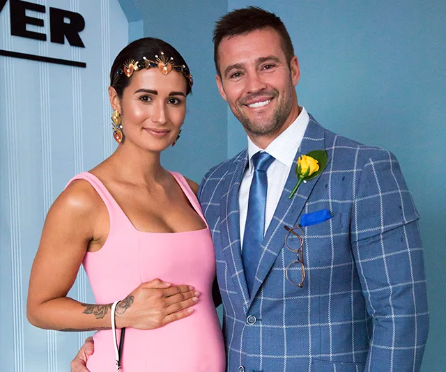 A couple poses together; the woman in a pink dress, cradling her belly, and the man in a blue checkered suit with a flower pin.