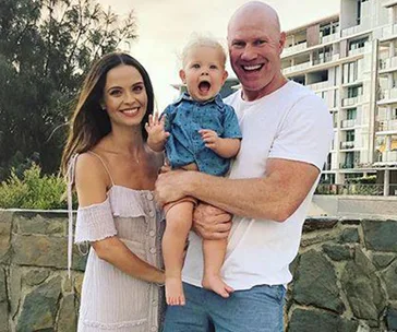 A happy family of three posing outdoors with a city building and trees in the background.