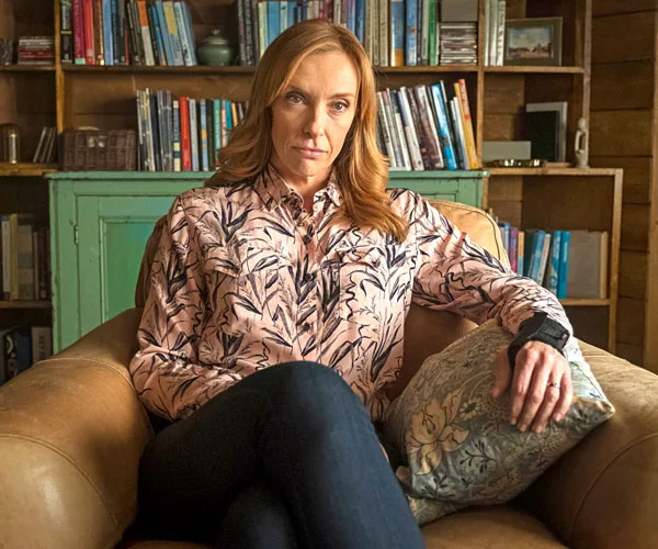 Toni Collette sitting in a chair, wearing a patterned shirt, in a cozy living room with bookshelves in the background.