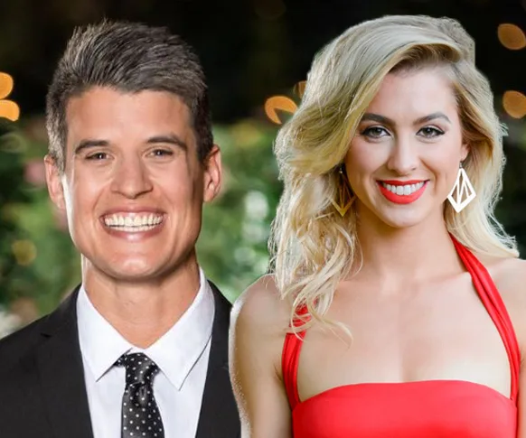 Two people smiling, one wearing a suit and tie, the other in a red dress, with blurred lights in the background.