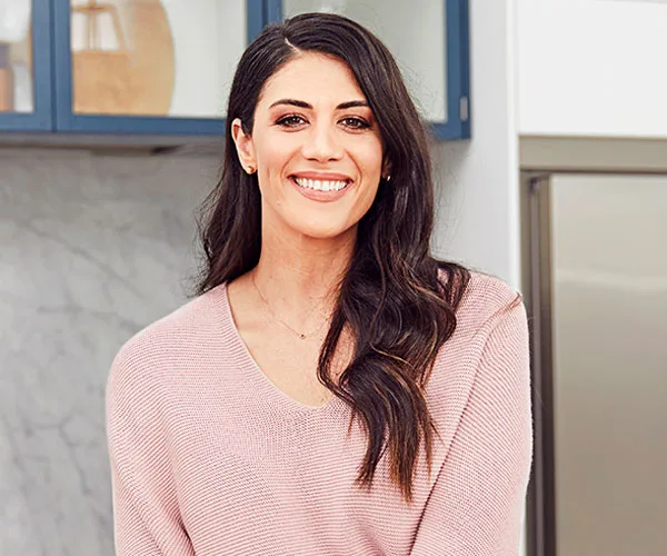 Smiling woman with long hair in a light sweater, standing in a modern kitchen setting.