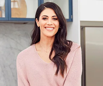 Smiling woman with long hair in a light sweater, standing in a modern kitchen setting.