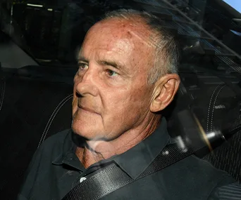 A man with short hair sits in a vehicle, wearing a dark shirt and seatbelt, looking out the window.