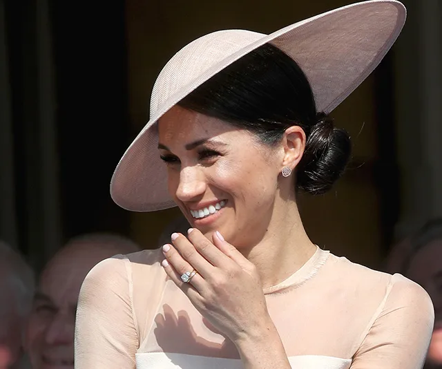 A woman smiling, wearing a light pink dress and matching wide-brimmed hat, with her hand raised to her face.