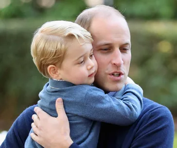 Father holding a young child closely, both in blue sweaters, in an outdoor setting with a blurred green background.