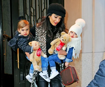 Woman holding toddler twins with teddy bears, wearing winter coats and hats, smiling outside a building.