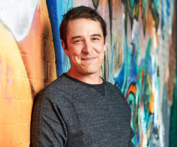 Man smiling, leaning against a graffiti-covered wall, wearing a grey sweater.