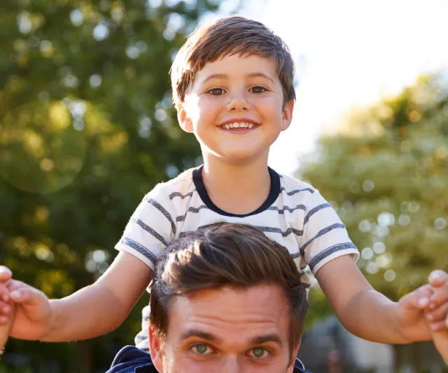 A smiling child on a man's shoulders in a park, with trees in the background.