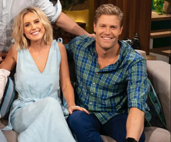 A man and woman sitting on a couch, smiling, alongside another partially visible person, indoors, dressed casually.