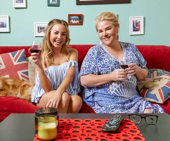 Two people sitting on a couch drinking wine, with a dog and British flag cushions in the background.