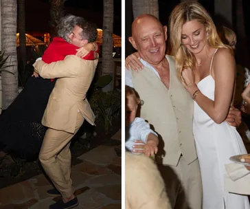 People celebrating at a wedding, one hugs joyfully while others smile and embrace, dressed in light attire.