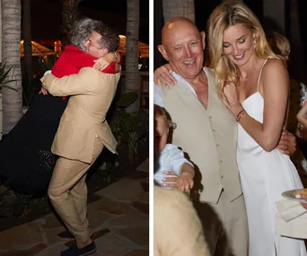 People celebrating at a wedding, one hugs joyfully while others smile and embrace, dressed in light attire.