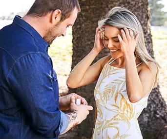 Man proposing to a woman with blonde hair in a white dress under a tree, both smiling and happy.