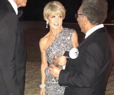 A woman in a glittering one-shoulder dress talks to two men in tuxedos at a formal event.