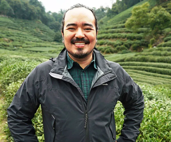 Man smiling in a tea plantation, wearing a black jacket and green checkered shirt, surrounded by lush greenery.
