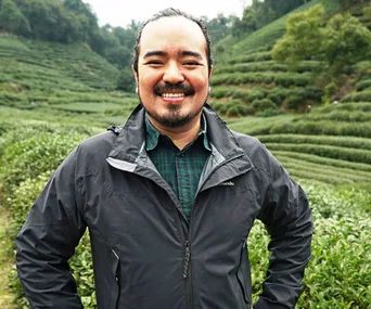 Man smiling in a tea plantation, wearing a black jacket and green checkered shirt, surrounded by lush greenery.