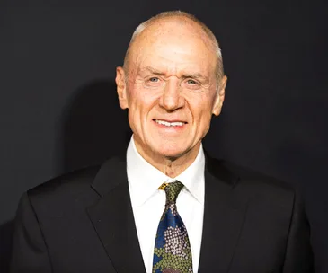 Man in a black suit with a colorful tie, smiling against a dark background.