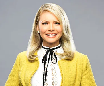 Portrait of a smiling woman with blonde hair, wearing a yellow jacket and a white blouse with a black ribbon tie.