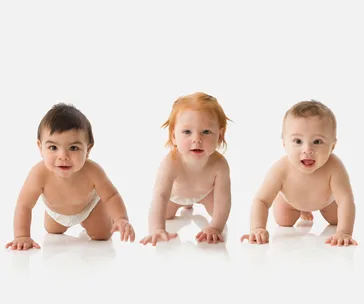 Three babies in diapers are crawling on a white surface, each smiling at the camera.