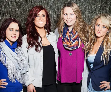 Four women smiling and posing together against a textured background; all wearing casual, colorful outfits.