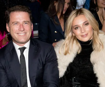 A smiling man in a suit and a woman in a fur-trimmed coat seated at an event, with a crowded background.