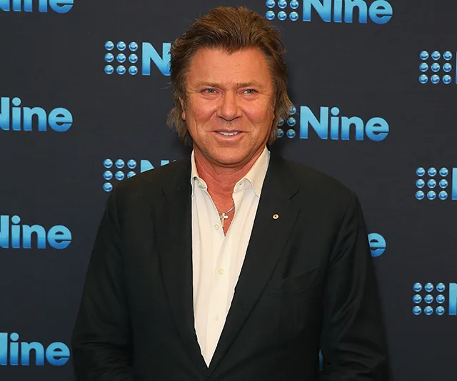 Man in a black suit with a white shirt stands smiling in front of a Nine Network backdrop.