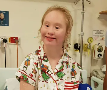 Girl in festive pajamas smiling while sitting in a hospital room with medical equipment in the background.