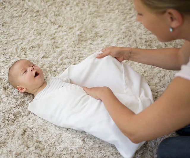 A parent gently swaddling a yawning baby in a white blanket on a soft carpet.