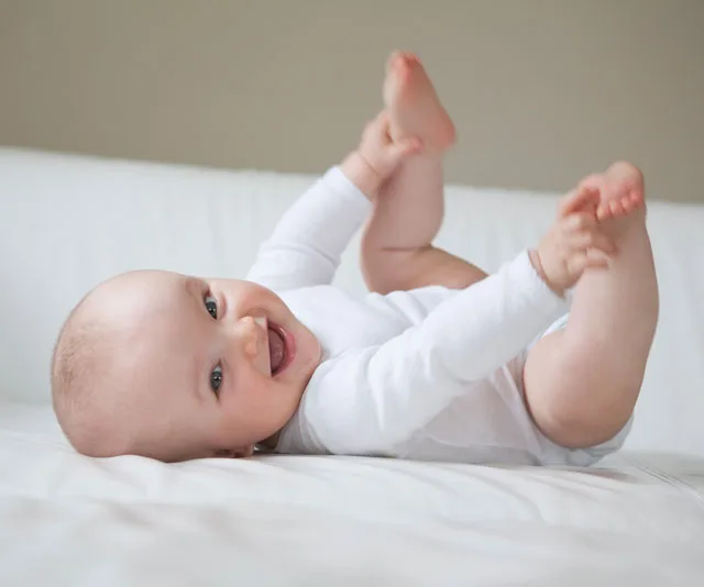 A baby in a white onesie lies on a bed, smiling and reaching for their toes.