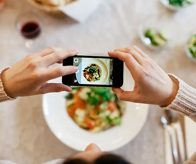 A person takes a photo of a colorful salad on a smartphone at a dining table.