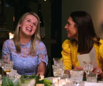 Two women seated at a dinner table, one in a blue lace top laughing, the other in yellow, holding a menu and smiling.
