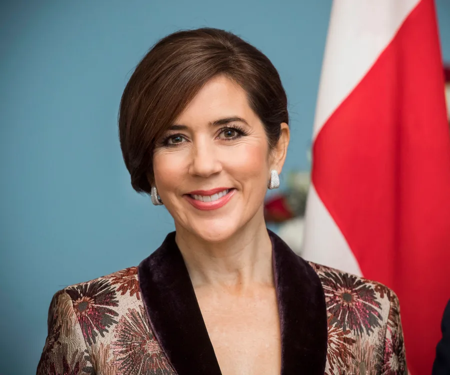 A woman with short brown hair, wearing a patterned jacket, smiling in front of a flag.