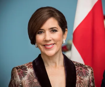 A woman with short brown hair, wearing a patterned jacket, smiling in front of a flag.
