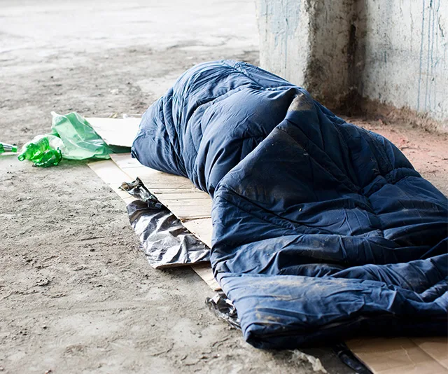 A person is lying in a blue sleeping bag on a cardboard in a rough concrete environment, with green plastic nearby.