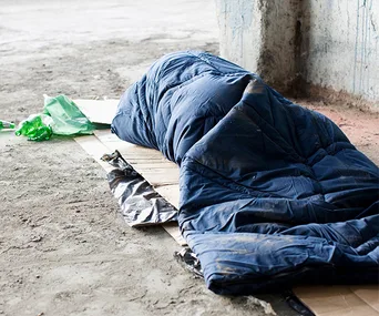 A person is lying in a blue sleeping bag on a cardboard in a rough concrete environment, with green plastic nearby.