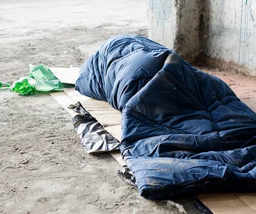 A person is lying in a blue sleeping bag on a cardboard in a rough concrete environment, with green plastic nearby.