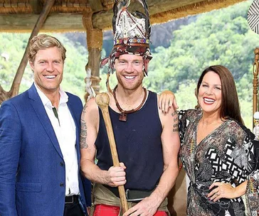 Three smiling people; the center person wears tribal attire with a headdress and holds a staff.