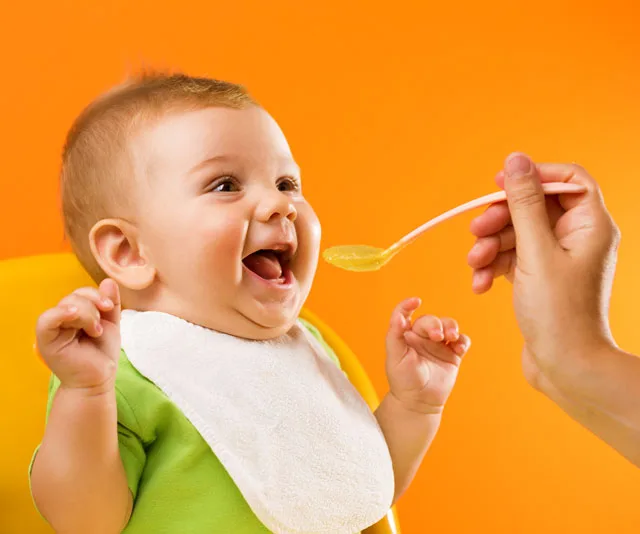 A happy baby in a green bib being fed with a spoon against a bright orange background.