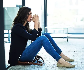 Woman sitting on floor, leaning against wall, looking pensive with hands clasped near mouth.