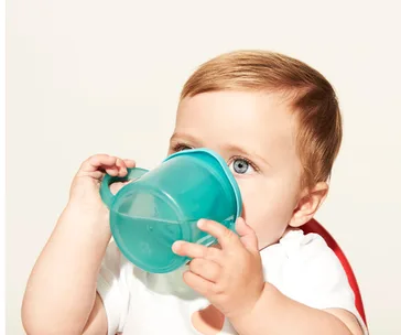 Baby drinking from a turquoise sippy cup, wearing a white shirt with a neutral background.