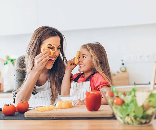 Mother and daughter in aprons playfully hold bell pepper slices in a kitchen with various vegetables on the counter.