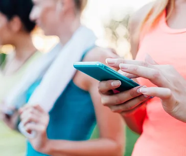 Women in athletic wear using a smartphone, possibly for a fitness app, with one holding a towel in the background.